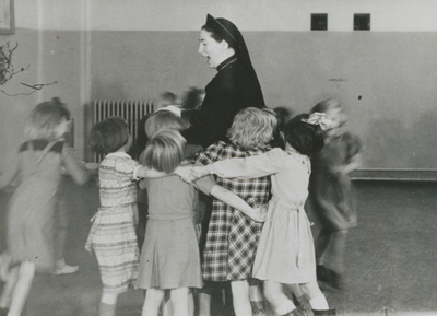 876947 Afbeelding van een Zuster Augustines die in haar functie als 'kleuterjuf' danst met een groep kleuters in de RK ...